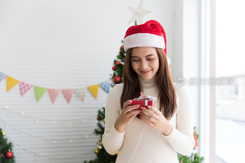 Smiling brunette woman in sweater and Santaâs hat holding small gift box and rejoices over Christmas decorative white background